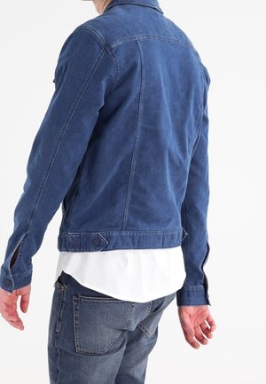 Mężczyzna w dopasowanej niebieskiej kurtce jeansowej na białej koszuli i niebieskich dżinsach, pokazany od tyłu na jednolitym białym tle.