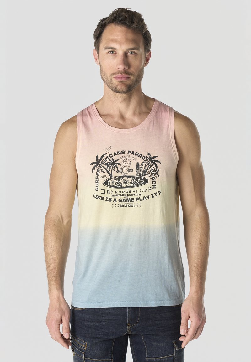 Hombre vistiendo camiseta sin mangas de tono pastel degradado con estampado negro de surf y texto, combinado con jeans oscuros, posando frente a un fondo claro.