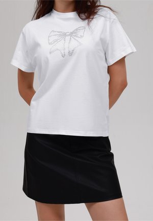 Frau trägt ein weißes T-Shirt mit gepunktetem Schleifendesign und einen schwarzen Rock, steht mit hinter dem Rücken verschränkten Händen vor einem einfarbigen Hintergrund.