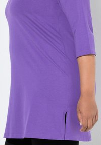 Fioletowa tunika z rękawami o długości do łokci, wykonana z miękkiego materiału. Posiada boczne rozporki u dołu dla ułatwienia ruchu.