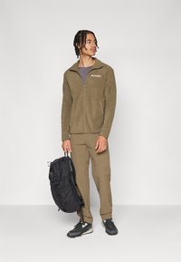 Fleece jacket in olive green with a zip front and logo. Paired with tan pants, black backpack, and dark shoes. Smooth texture, casual design.