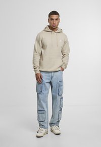 Beige hættejakke med en frontlomme og logo, parret med lyseblå denim cargo-bukser med flere sidelommer, komplementeret af hvide sneakers.