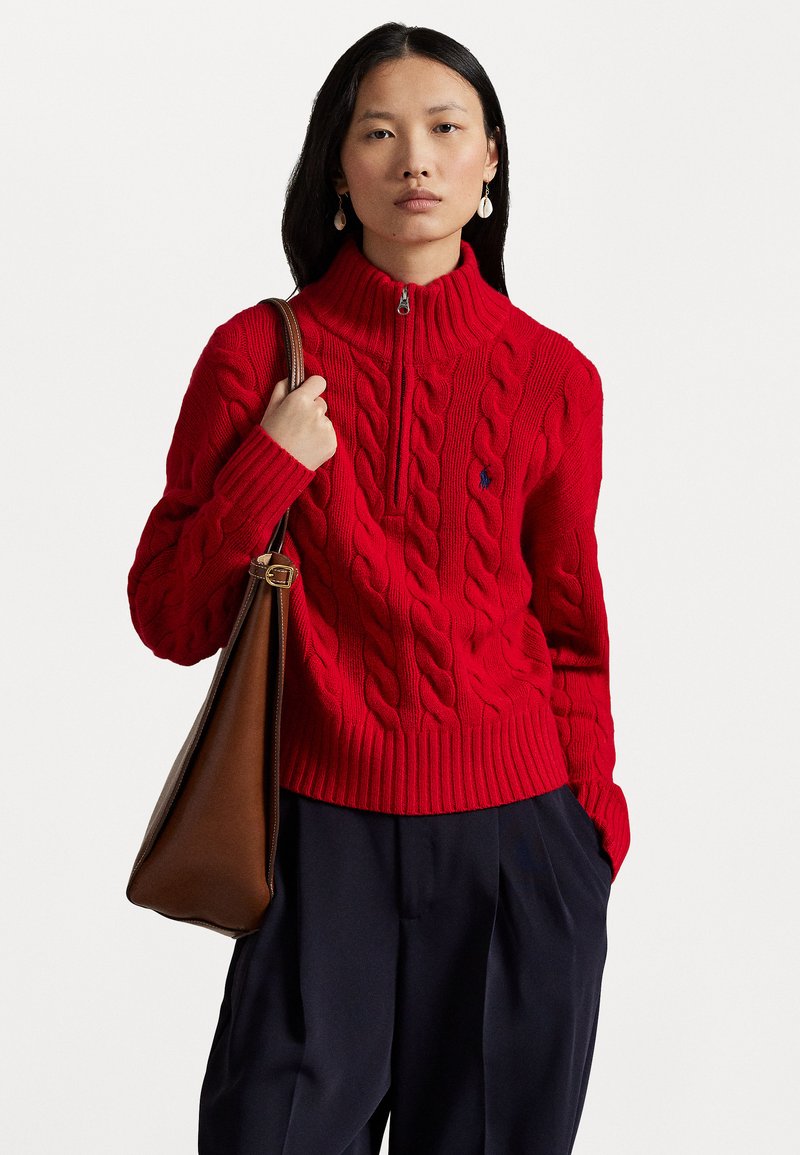 Red knitted sweater with a high collar, featuring cable knit patterns. Paired with dark trousers and a brown handbag. Zipper detail at neck.