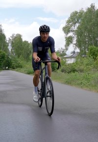 Dviračių entuziastas su juodu kelių dviračiu, dėvintis prigludusią tamsią maikutę, juodą šalmą ir baltus batus, važiuoja medžiais apsodintu taku.
