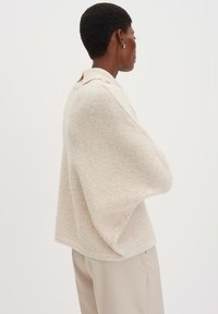 Pullover beige, con texture e colletto, caratterizzato da maniche larghe e una vestibilità rilassata. Indossato sopra pantaloni su misura di colore chiaro.