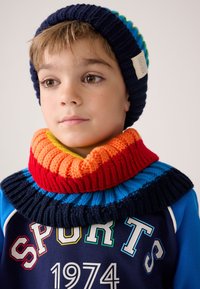 Pletená čepice v námořnické modři s pruhovaným vzorem v modré, zelené a korálové barvě; žebrovaný nákrčník v červené, oranžové a žluté barvě s texturovaným povrchem.