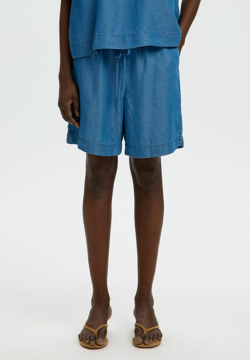 Personne portant un short en jean bleu ample avec cordon de serrage et un haut assorti, debout pieds nus dans des sandales tongs beige sur fond blanc.
