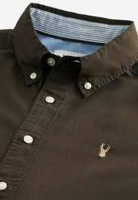 Chemise marron boutonnée avec col rayé contrastant. Comprend cinq boutons blancs et un petit cerf brodé sur la poitrine. Texture lisse.