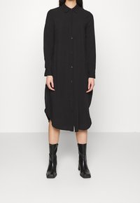 Robe chemise noire à boutons, avec un col, des manches longues et des fentes latérales ; tissu lisse avec une coupe décontractée, associée à des bottes noires montantes.