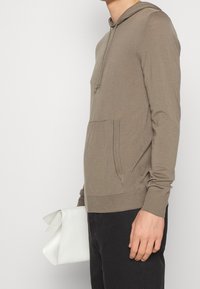 Beige munkjacka med känguruficka framtill, ribbade ärmslut och nederkant, bärs med en vit kuvertväska. Slät tygtextur.