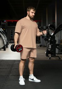 T-shirt e pantaloni sportivi beige con vestibilità rilassata, che tiene un manubrio nero con un'etichetta di peso rossa e nera. Sneakers bianche con dettagli neri.