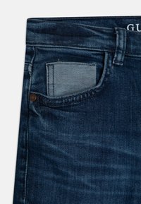 Ciemnoniebieskie jeansy z przednią kieszenią ozdobioną jaśniejszym materiałem dżinsowym. Faktura jest gładka z delikatnymi efektami przetarć.