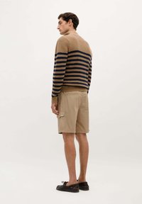 Striktrøje i tan med marineblå vandrette striber, parret med beige cargo shorts. Modellen bærer mørke sko og står foran en neutral baggrund.