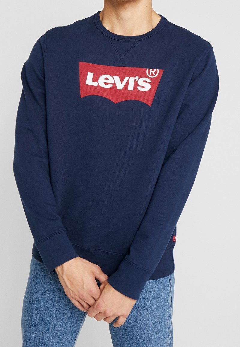 Persona vistiendo una sudadera Levi's azul marino con el logo en rojo y blanco, combinada con vaqueros azul claro, con las manos entrelazadas al frente.