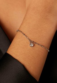 Zilveren kettingarmband met een kleine ronde hanger met een heldere steen, rustend op een gebruinde pols tegen een donkere achtergrond.