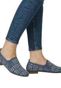 Blaue Slipper aus strukturiertem Stoff, mit einem geometrischen Silbermuster, flachem Absatz und runder Zehenpartie, getragen mit dunklen Jeans.