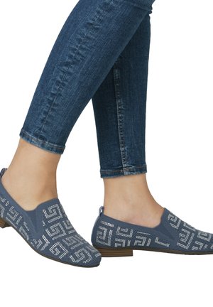 Blue slip-on shoes made of textured fabric, featuring a geometric silver pattern, low heel, and rounded toe, worn with dark denim jeans.