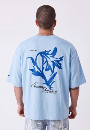 Uomo con capelli corti che indossa una maglietta azzurro chiaro con un grande motivo floreale blu e il testo "Change the Game" sulla schiena.