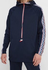 Mörkblå zip-hoodie med huva och kenguruficka. Har en röd dragkedja, vita och blå sidostripes samt en liten röd Nike-logotyp på framsidan.