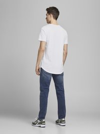 Camiseta blanca de manga corta, jeans azules y zapatillas verdes y blancas. Costuras visibles y ligeros desgastes en los jeans. Vista trasera.