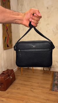 Hand houdt zwarte leren BOSS polstas bij de band tegen een wazige binnenachtergrond met houten vloer.