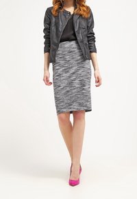 Veste en cuir noire, haut noir ajusté, jupe crayon grise texturée et escarpins rose vif. Le mannequin pose devant un fond blanc.