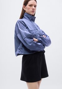 Ελαφρύ, κομμένο σακάκι από ματ ύφασμα λεβάντας με ψηλό γιακά και ελαστικά μανίκια, συνδυασμένο με μαύρα σορτς με κορδόνι στη μέση.