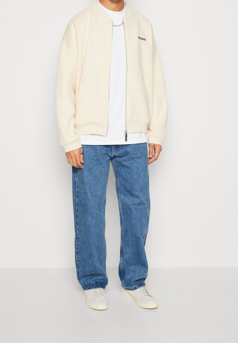 Personne portant une veste zippée crème par-dessus une chemise blanche, un jean bleu ample, des baskets blanches et un collier chaîne argenté sur un fond uni.