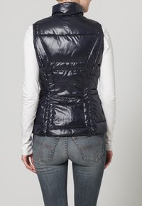 Mujer con un chaleco acolchado azul marino oscuro sobre una camiseta de manga larga blanca y jeans azules, vista desde atrás.