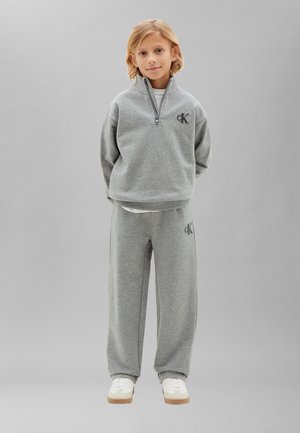 Enfant aux cheveux blonds debout, portant un survêtement gris Calvin Klein et des baskets blanches sur un fond gris clair uni.