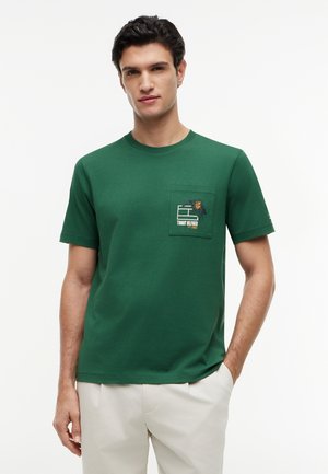 Mann trägt grünes kurzärmeliges T-Shirt mit Brusttasche, verziert mit floralem Muster und weißem Tommy Hilfiger Logo, kombiniert mit weißer Hose.