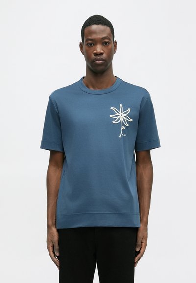 T-shirt azul de algodão com mangas curtas, apresentando um design de flor bordado em branco do lado esquerdo e uma bainha reta.