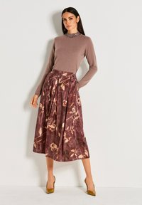 Vrouw in mauve langekranslang shirt met een pailletten kraag, een bloemenpatroon geplisseerde midi rok en mosterdkleurige hoge hakken, staand tegen een witte muur.