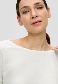 Weißes Leinenoberteil mit weitem Ausschnitt, weicher Textur und lässigem Schnitt, getragen von einer Person vor neutralem Hintergrund.
