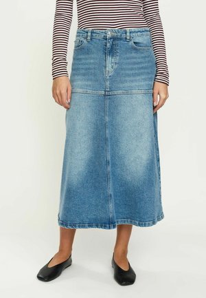 Persona che indossa una gonna midi di jeans blu, ballerine nere e una maglia a maniche lunghe a righe marroni e bianche, in piedi davanti a uno sfondo semplice.