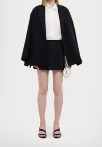 Yli suuri musta bleiseri valkoisen kauluspuseron päällä, musta laskostettu mini-hame ja mustat kiilanauhalaput. Valkoinen käsilaukku kädessä.