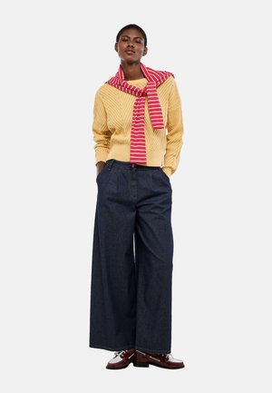 Femme debout, mains dans les poches, portant un pull jaune, une écharpe rayée rouge et blanche, un jean large foncé et des chaussures en cuir bicolores.