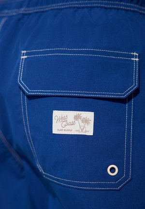 Kék fürdőnadrág hátsó zsebbel, amelyen egy fehér címke található, rajta a "West Coast Surf Supply" felirattal, fehér varrással és egy szemmel.