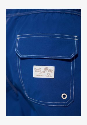 Shorts de bain bleu avec une poche arrière portant une étiquette blanche affichant "West Coast Surf Supply", accentués par des coutures blanches et un œillet.