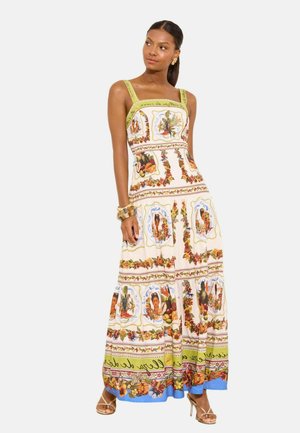 Femme portant une longue robe sans manches avec des motifs floraux et fruités colorés, des bretelles vertes et des sandales dorées à talons hauts.