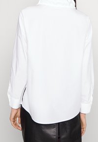 Camicia bianca a maniche lunghe con colletto alto, dotata di polsini con bottoni e orlo curved. Tessuto morbido e leggero con una texture liscia.