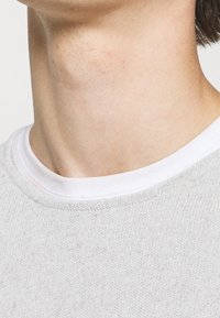 Pull tricot gris clair avec un col côtelé blanc. Le tissu est texturé avec un motif subtil, mettant en valeur son design en tricot doux.