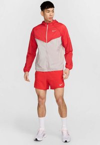 Veste coupe-vent Nike rouge et grise avec zip sur le devant, capuche et motif en chevron ; associée à un short rouge et des chaussettes blanches. Baskets blanches.