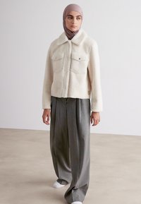 Weißer Kunstfaserpelzjacke mit Kragen, zwei Brusttaschen und strukturiertem Stoff, kombiniert mit grauen weit geschnittenen Hosen und weißen Schuhen.