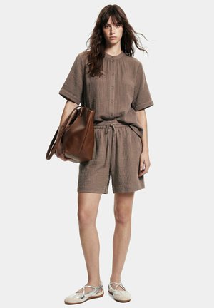 Donna in piedi indossa una camicia marrone ampia abbottonata e shorts coordinati, tiene una grande borsa marrone e indossa scarpe basse bianche.
