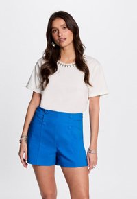 Witte top met korte mouwen en een decoratieve halslijn, gecombineerd met helderblauwe high-waisted shorts met knoopaccenten en zijzakken.