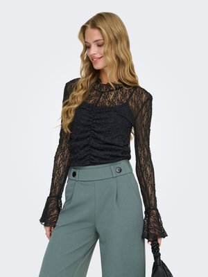Woman in long-sleeve black lace top and high-waisted green trousers holding a black handbag, looking down and smiling.
