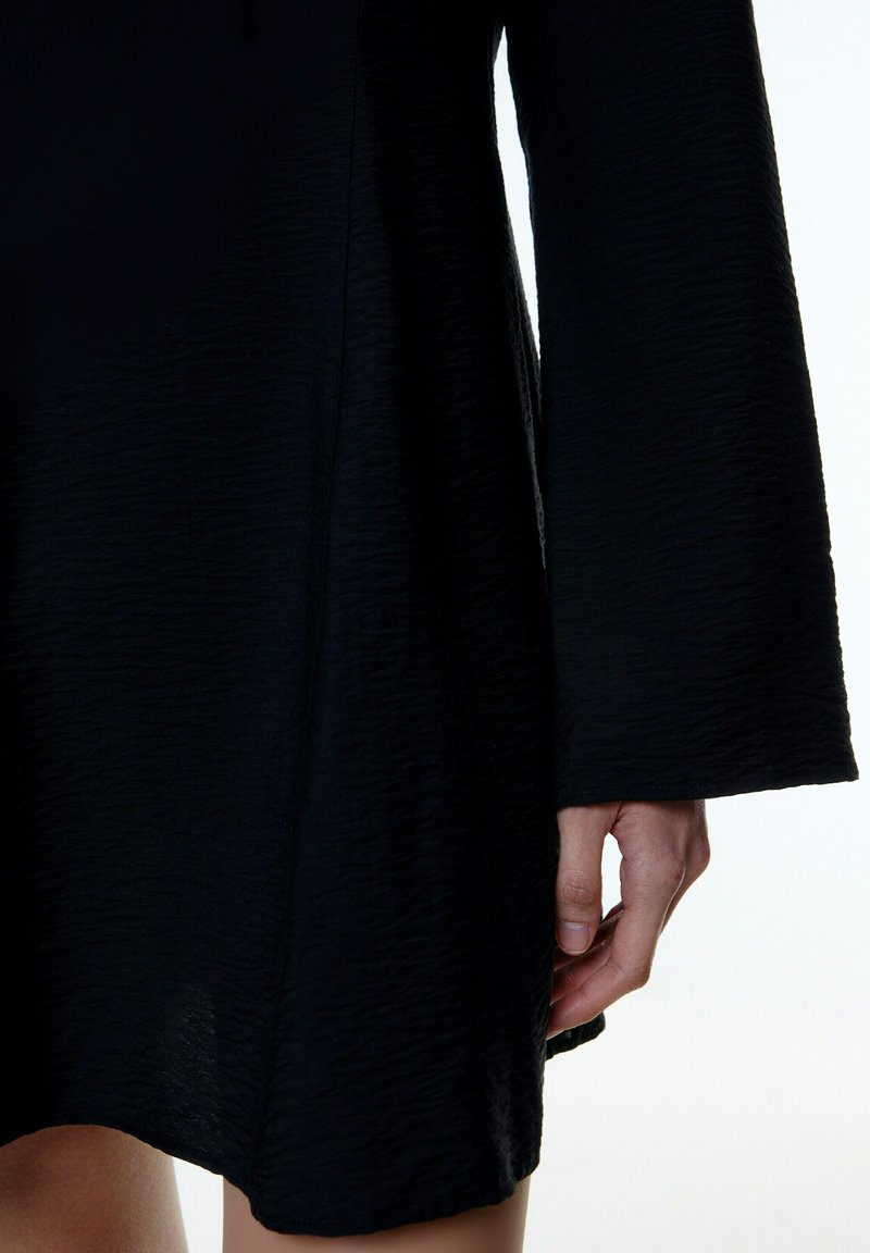 Schwarzes Kleid aus einem strukturierten, zerknitterten Stoff. Mit langen Ärmeln und einem lockeren Schnitt. Seitliche Nahtdetails entlang des Saums sichtbar.