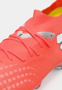 Zapatilla de fútbol de color rojo brillante con superficie texturizada, diseño elegante, detalles en plata y sistema de cordones tradicional, con un logotipo prominente.