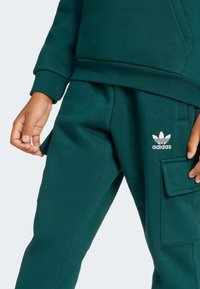Pantalons de jogging en coton verts avec une taille côtelée, une poche latérale et un logo Adidas blanc sur la cuisse. Texture lisse et design ajusté.